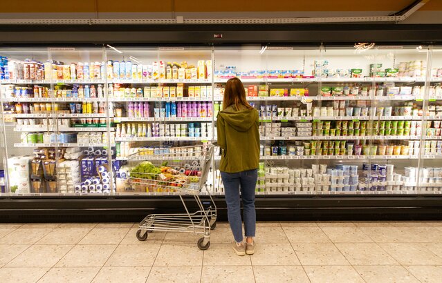 Blonde Frau steht mit halbvollem Wagerl vor Milchprodukten im Supermarkt | © Land schafft Leben